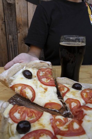 Photo for Closeup view of a woman having a black beer and a slice of pizza with cheese, tomatoes and black olives. - Royalty Free Image
