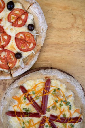 Téléchargez les photos : Vue de dessus de deux pizzas sur la table en bois. Une avec du fromage mozzarella, des tranches de tomate et des olives noires, et l'autre avec de la mozzarella, du cheddar, de l'oignon vert et des saucisses fumées. - en image libre de droit