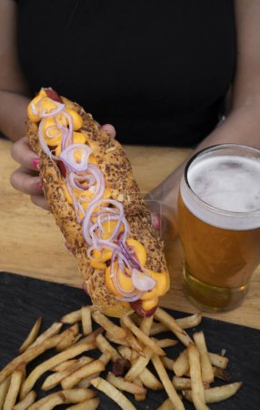 Photo for Gourmet fast food. Closeup view of a woman eating a hot dog with red onion and cheddar cheese, with french fries and a glass of beer. - Royalty Free Image