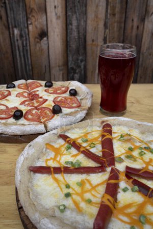 Photo for Closeup view of a pizza with mozzarella and cheddar cheese, green onion and smoked sausages, and a pizza with sliced tomatoes and black olives, with a pint of beer. - Royalty Free Image