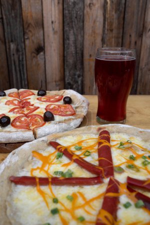 Foto de Vista de cerca de una pizza con mozzarella y queso cheddar, cebolla verde y salchichas ahumadas, y una pizza con tomates en rodajas y aceitunas negras, con una pinta de cerveza. - Imagen libre de derechos