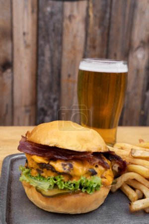 Photo for Hamburger. Closeup view of a burger with grilled meat, lettuce, cucumber pickles, cheddar cheese and crispy bacon with a glass of beer and french fries. - Royalty Free Image