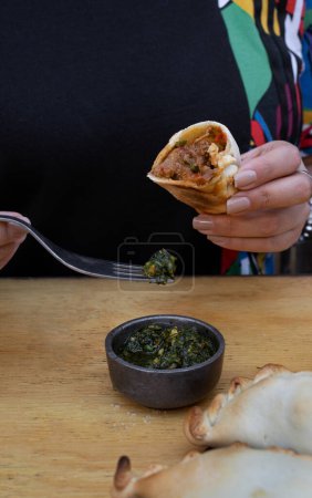 Photo for Eating empanadas at the restaurant. Closeup view of a woman hands pouring spicy chimichurri sauce with a spoon into a meat empanada. - Royalty Free Image