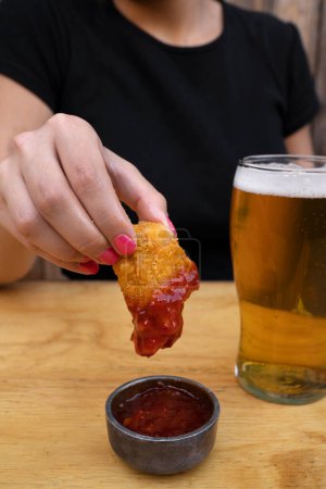 Photo for Gastronomy. Closeup view of a woman holding a fried mozzarella cheese stick dipped in a spicy sauce, and having a beer. - Royalty Free Image