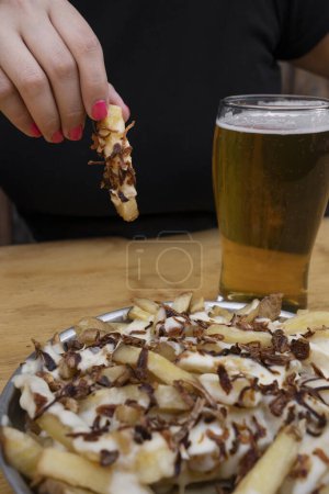 Foto de Papas fritas. Vista de cerca de una mujer comiendo papas fritas con queso derretido y cebolla crujiente, y tomando una pinta de cerveza. - Imagen libre de derechos