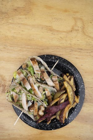 Foto de Gastronomía de fusión. Vista superior de un plato negro con tacos de ensalada César y batatas fritas en la mesa de madera. - Imagen libre de derechos