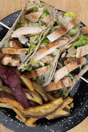 Photo for Fusion gastronomy. Top view of a black dish with Caesar salad tacos and fried sweet potatoes on the wooden table. - Royalty Free Image