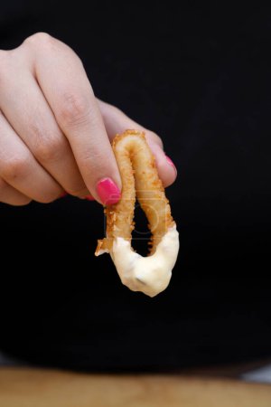 Foto de Comer mariscos. Vista de cerca de una mujer sosteniendo un anillo de calamar frito sumergido en salsa aioli. - Imagen libre de derechos