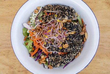 Photo for Healthy food. Top view of a salad with carrot, lettuce, peanuts, chicken breast breaded with seeds, soy sprouts and cabbage, in a white bowl on the wooden table. - Royalty Free Image
