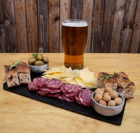 Foto de Picada. Vista de cerca de un plato con salami en rodajas, queso, cacahuetes focaccia, aceitunas verdes y una pinta de cerveza en la mesa de madera. - Imagen libre de derechos