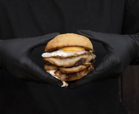 Téléchargez les photos : Chef portant des gants tenant un hamburger monstre avec viande, fromage provolone, bacon, sauce barbecue, rondelles d'oignons et un œuf grillé, avec un fond noir. - en image libre de droit
