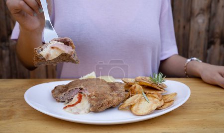 Foto de Mujer sosteniendo un tenedor, tomando una milanesa, bistec frito empanado, relleno con queso mozzarella, jamón y pimiento morrón con patatas fritas y limón, en un plato blanco. - Imagen libre de derechos