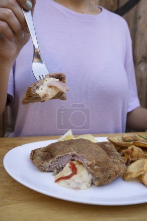 Photo for Woman holding a fork, having a milanesa, breaded fried steak, stuffed with mozzarella cheese, ham and bell pepper with Spanish chips and lemon, in a white dish. - Royalty Free Image
