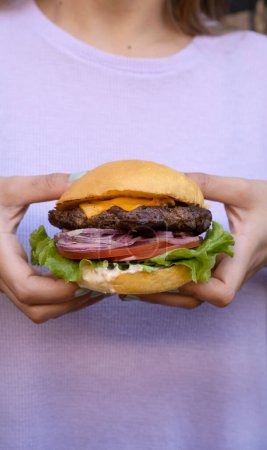Téléchargez les photos : Présentation du hamburger. Vue rapprochée d'une femme tenant un hamburger avec laitue, tomate, fromage cheddar, oignon rouge et mayonnaise. - en image libre de droit