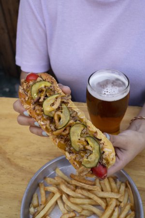 Photo for Gourmet fast food. Closeup view of a woman eating a hot dog with crispy onion, sliced cucumber and mustard with honey, with french fries and a glass of beer. - Royalty Free Image