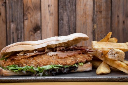 Foto de Sandwich gourmet. Vista de cerca de un sándwich multicapa con filete frito de pollo, tocino, queso y lechuga, con papas fritas. - Imagen libre de derechos
