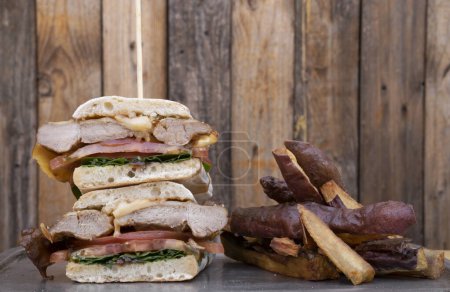 Téléchargez les photos : Sandwich gourmand. Vue rapprochée d'un sandwich multicouche avec pain ciabatta, filet de porc grillé, fromage, roquette et oignon, dans un plat en métal avec patates douces frites, avec un fond en bois. - en image libre de droit