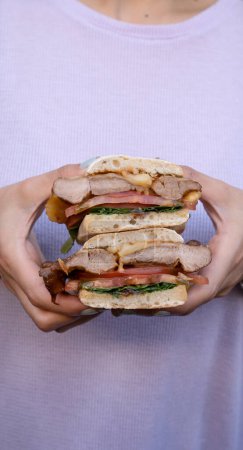 Téléchargez les photos : Sandwich gourmand. Vue rapprochée de la femme tenant un sandwich multicouche avec pain ciabatta, bondiola, fromage, roquette et oignon. - en image libre de droit