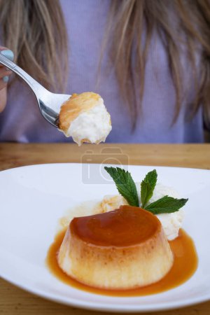 Photo for Eating dessert. Closeup view of a woman's hand holding a spoon, having a flan with cream and caramel, in a white dish on the restaurant wooden table. - Royalty Free Image