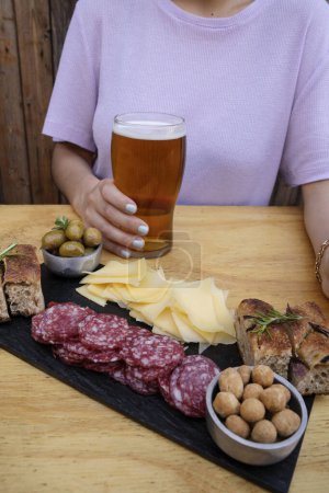 Foto de Picada. Vista de cerca de una mujer bebiendo una cerveza y tomando embutidos, como salami en rodajas, queso, cacahuetes focaccia y aceitunas verdes. - Imagen libre de derechos