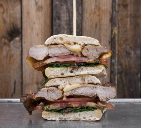 Foto de Sandwich gourmet. Vista de cerca de un sándwich multicapa con pan de ciabatta, lomo de cerdo a la parrilla, queso, rúcula y cebolla, en un plato de metal, con un fondo de madera. - Imagen libre de derechos