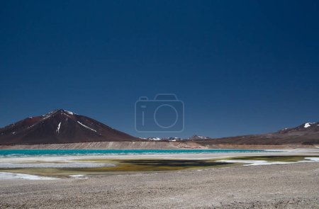 Foto de Paisaje volcánico. Vista del estanque amarillo y negro debido al azufre en el agua, lago de agua glaciar turquesa, llano de sal natural y montañas de los Andes en el fondo, bajo un cielo azul profundo. - Imagen libre de derechos