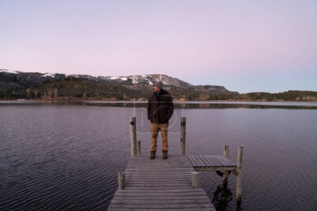 Téléchargez les photos : Le lac placide à la tombée de la nuit. Vue magique d'un homme debout dans le quai en bois avec les montagnes et la forêt en arrière-plan. Belles couleurs de coucher de soleil dans le ciel et l'eau. - en image libre de droit