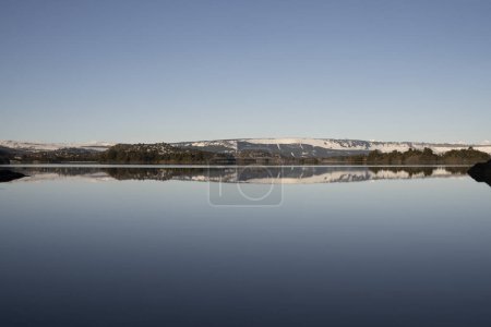 Téléchargez les photos : Symétrie dans la nature. Vue sur le lac calme et le rivage à l'horizon. La forêt et le ciel bleu clair se reflètent à la surface de l'eau. - en image libre de droit