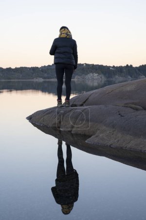 Foto de Retrato de una joven de pie en la playa rocosa contemplando el lago, el bosque y las montañas al atardecer. Su reflejo perfecto en el agua. - Imagen libre de derechos