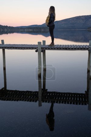 Téléchargez les photos : Portrait magique d'une jeune femme debout sur le quai en bois dans le lac calme au coucher du soleil. Sa silhouette parfaite se reflète dans l'eau. Les montagnes, la forêt, le rivage et l'horizon en arrière-plan. - en image libre de droit