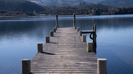 Téléchargez les photos : Les quais en bois dans le lac calme. Le ciel bleu clair et la jetée se reflètent dans la surface de l'eau. La forêt et les montagnes en arrière-plan. - en image libre de droit