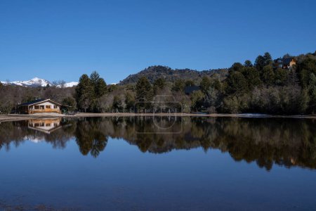 Foto de La casa del lago en un día soleado. Vista de la cabaña, bosque, montañas y reflejo del cielo azul en la superficie del agua. - Imagen libre de derechos