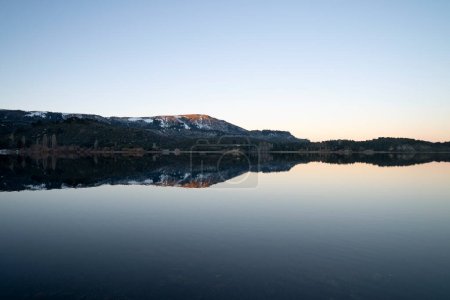 Téléchargez les photos : Vue enchanteresse sur le lac placide au coucher du soleil. Les montagnes, la forêt et le ciel bleu se reflètent dans l'eau. - en image libre de droit