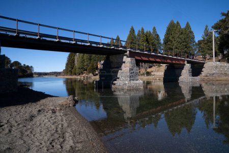 Téléchargez les photos : Le pont traversant la rivière reliant deux lacs. Sa réflexion à la surface de l'eau. Le rivage de la plage et la forêt dans une journée d'été ensoleillée avec un ciel bleu clair. - en image libre de droit