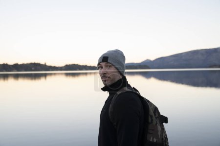 Photo for Portrait of a caucasian man in his 30s, wearing a hat, scarf and jacket with the lake and mountains in the background, at sunset. Beautiful landscape and sky reflection in the water surface. - Royalty Free Image