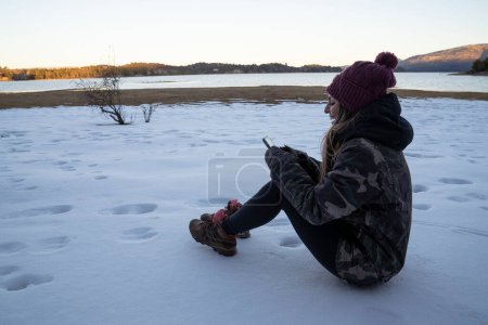 Téléchargez les photos : Portrait d'une jeune femme dans la vingtaine, vêtue d'un chapeau et d'une veste, assise dans la neige et jouant avec le téléphone portable. Le lac et les montagnes au coucher du soleil en arrière-plan. - en image libre de droit
