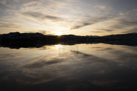 Foto de Puesta de sol mágica en el lago. Vista panorámica del lago de almejas, sol escondido y cielo dramático con nubes, hermoso reflejo en la superficie del agua. El bosque y las montañas en el horizonte. - Imagen libre de derechos