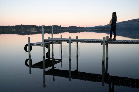Téléchargez les photos : Silhouette d'une jeune femme debout sur le quai en bois à la tombée de la nuit. Vue magique sur le lac, les montagnes et la forêt et son reflet dans l'eau du lac. Belles couleurs de coucher de soleil. - en image libre de droit