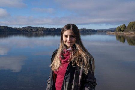 Téléchargez les photos : Voyage. Portrait d'une jeune femme caucasienne adulte dans la vingtaine, souriante, avec le lac paisible en arrière-plan. Les montagnes, la forêt et le ciel bleu se reflètent dans l'eau. - en image libre de droit