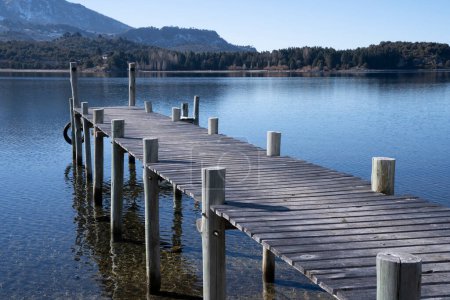 Téléchargez les photos : Le quai en bois dans le lac calme. Le ciel bleu clair et la jetée se reflètent dans la surface de l'eau. La forêt et les montagnes en arrière-plan. - en image libre de droit