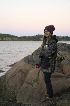 Photo for Portrait of a young woman in her 20s, wearing a hat and a jacket, with the lake in the background, at sunset. - Royalty Free Image