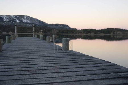Téléchargez les photos : Concentration sélective sur les bois de la jetée dans le lac. Les Alpes et la forêt se reflètent dans l'eau calme du lac dans le fond. Belles couleurs de coucher de soleil. - en image libre de droit
