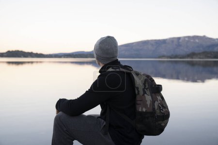 Photo for Tourism. Portrait of a young man wearing a hat and bag, contemplating the beautiful landscape, lake and mountains, at sunset. - Royalty Free Image