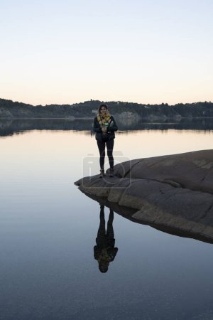 Téléchargez les photos : Portrait d'une jeune femme debout sur le rivage rocheux et reflet de sa silhouette dans le lac. La forêt et les collines en arrière-plan. Belles couleurs de coucher de soleil dans l'environnement. - en image libre de droit