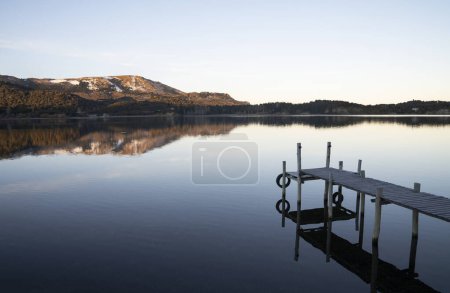 Téléchargez les photos : Vue magique sur le quai en bois dans le lac placide au lever du soleil. Les montagnes, la forêt et le ciel bleu se reflètent dans l'eau. - en image libre de droit