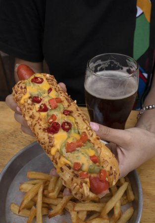 Téléchargez les photos : De la nourriture américaine. Vue rapprochée d'une femme tenant un hot-dog gastronomique avec guacamole, chilis et fromage cheddar. Des frites et une bière sur la table. - en image libre de droit