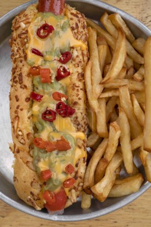Photo for Gourmet fast food. Top view of a hot dog with guacamole, chilis, cheddar cheese and french fries, in a metal dish with a wooden background. - Royalty Free Image