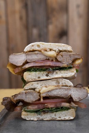 Foto de Sandwich gourmet. Vista de cerca de un sándwich multicapa con pan de ciabatta, bondiola, queso, rúcula y cebolla, en un plato de metal, con un fondo de madera. - Imagen libre de derechos