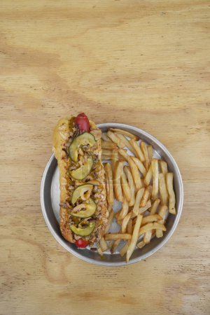 Foto de Comida rápida gourmet. Vista de cerca de un hot dog con pepino en rodajas, miel con mostaza y cebolla crujiente, con papas fritas en un plato de metal sobre la mesa de madera. - Imagen libre de derechos