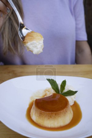 Photo for Eating dessert. Closeup view of a woman's hand holding a spoon, having a flan with cream and caramel, in a white dish on the restaurant wooden table. - Royalty Free Image